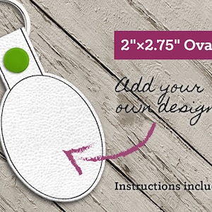 Puede incluir: Llavero ovalado de cuero blanco con un botón verde y un contorno negro. El llavero mide 5,08 cm por 7,0 cm. El texto "¡Añade tu propio diseño!" está escrito en rosa sobre un fondo de madera. Se incluyen instrucciones.
