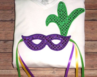 May include: A white t-shirt with a purple and green Mardi Gras mask with sequins and a green feather. The mask has purple, yellow, and green ribbons attached.