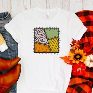 Könnte beinhalten: Weißes T-Shirt mit einem Patchwork-Design in Grün-, Gelb-, Orange- und Lilatönen. Das Design ist schwarz umrandet und hat einen genähten Look. Das Shirt liegt auf einer Holzoberfläche mit einem Paar braunen Stiefeln, einer Jeansjacke und einem karierten Schal.
