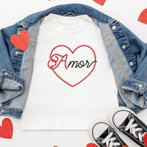 Peut inclure: Un t-shirt blanc avec un contour de cœur rouge et le mot "Amor" écrit en noir à l'intérieur du cœur. Le t-shirt est posé sur une surface blanche avec une veste en jean, des cœurs rouges et des baskets noires.