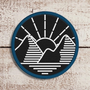 Puede incluir: Un parche redondo bordado con un diseño de paisaje montañoso con un sol naciente. El parche tiene un fondo negro con detalles blancos y un borde azul. El diseño incluye montañas estilizadas y rayos de sol.