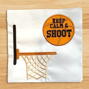 May include: Embroidered design of a basketball hoop with a basketball above it. The basketball has the text "Keep Calm & Shoot" on it.