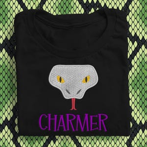 May include: Black t-shirt with a snake head design, yellow eyes, and a red tongue. The word "CHARMER" is embroidered in purple below the snake. The shirt is displayed on a green and black snake skin pattern.