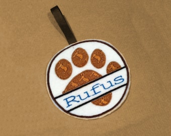 May include: A white round ornament with a brown paw print design and the name "Rufus" written in blue lettering. The ornament has a brown ribbon attached to the top.
