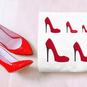 May include: A white t-shirt with red embroidered high heel shoes of different sizes. The shirt is lying flat on a wooden surface. Two red high heel shoes are also lying on the surface.