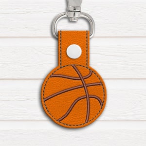 Puede incluir: Un llavero de cuero marrón con un diseño de baloncesto cosido. El llavero tiene un anillo de metal plateado y un botón blanco.