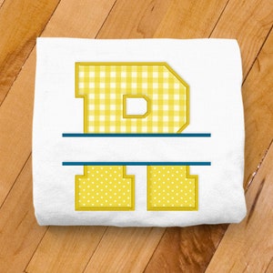 May include: A white t-shirt with a yellow letter "D" in a gingham pattern. The letter is outlined in yellow and has a blue line above and below it.