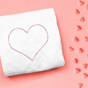 Puede incluir: Un cuadrado de tela blanca con un diseño de corazón cosido en rojo sobre un fondo rosa. Hay corazones rosas dispersos en el lado derecho de la imagen.