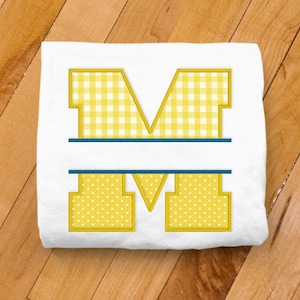 May include: A white t-shirt with a yellow and white gingham letter M with a blue line across the middle. The letter is outlined in yellow.