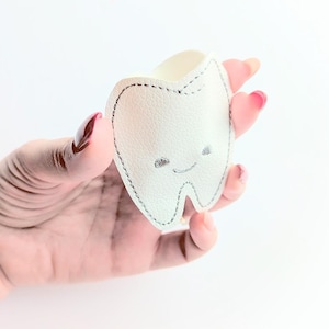 May include: A white, tooth-shaped pouch with a smiling face and gray stitching. The pouch is held in a hand against a white background. The pouch is made of a soft material.