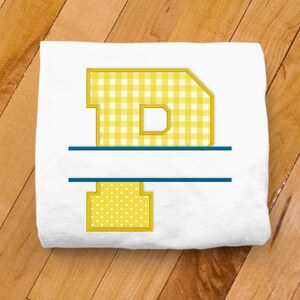 May include: A white folded towel with a yellow letter "D" design. The letter is divided by a horizontal blue line. The top portion of the letter has a yellow and white gingham pattern, while the bottom has yellow polka dots.