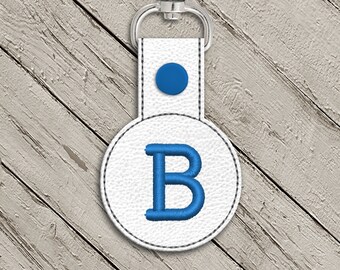 May include: White leather keychain with a blue embroidered letter "B" on a round tag. The keychain has a silver metal ring.
