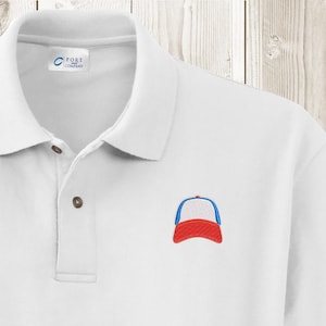 May include: White polo shirt featuring a small embroidered baseball cap design in red, white, and blue. The shirt has a classic collar, button placket, and the "Port Company" logo on the label.
