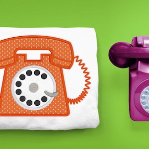 May include: A white t-shirt with an orange embroidered rotary phone design. The phone has white polka dots and a black dial. A real, purple rotary phone is shown to the right of the t-shirt.