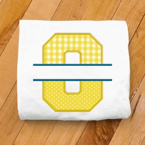 May include: A white onesie with a yellow number "0" in a split monogram style. The top half of the number is a yellow and white gingham pattern and the bottom half is a yellow and white polka dot pattern. The split monogram has a navy blue line across the middle.