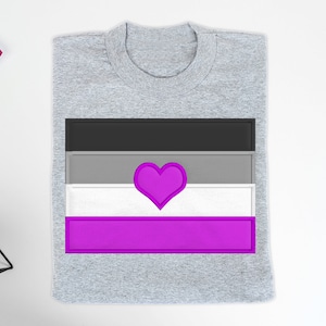 Könnte beinhalten: Ein graues T-Shirt mit einer gestickten asexuellen Pride-Flagge. Die Flagge hat schwarze, graue, weiße und violette Streifen mit einem violetten Herzen in der Mitte.