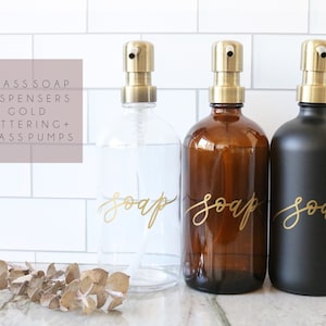May include: Three glass soap dispensers with gold lettering and brass pumps. The dispensers are clear, amber, and black. The dispensers are labeled "soap" in gold lettering.