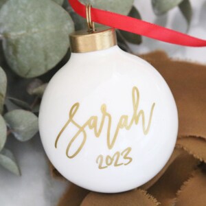 May include: White ceramic Christmas ornament with gold lettering that reads "Sarah 2023".