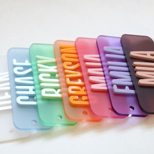 May include: A collection of colorful name tags in a row. Each tag is a different color, including clear, blue, green, orange, pink, purple, and brown. Each tag has a name in a contrasting color, such as white or light brown.