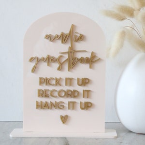 May include: A light pink acrylic sign with gold lettering that reads "audio guestbook." Below, it says "Pick it up, Record it, Hang it up." A small gold heart is at the bottom. A white vase with dried flowers is in the background.