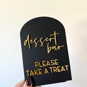May include: An arched black sign featuring gold 3D lettering. The top portion displays the words "dessert bar" in a cursive script, while the bottom section features the text "PLEASE TAKE A TREAT" in a bold, capitalized sans-serif font.