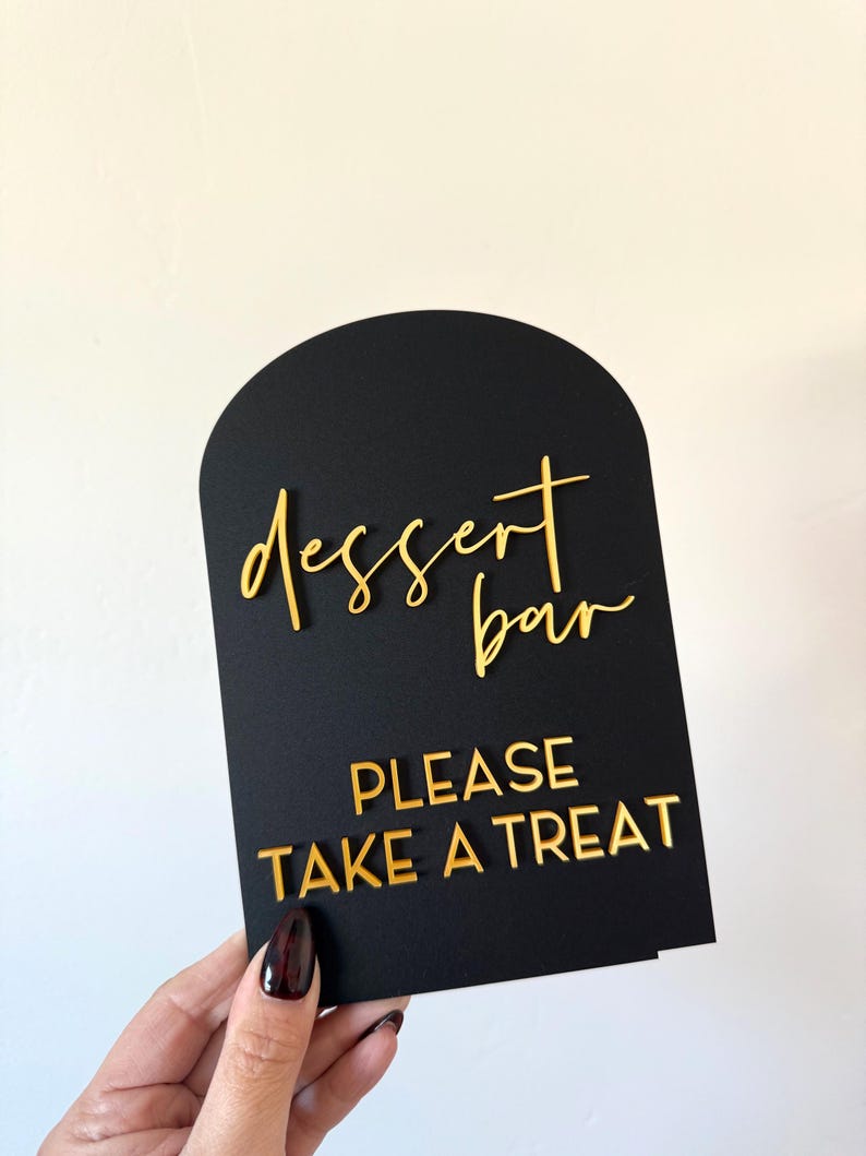 May include: An arched black sign featuring gold 3D lettering. The top portion displays the words "dessert bar" in a cursive script, while the bottom section features the text "PLEASE TAKE A TREAT" in a bold, capitalized sans-serif font.