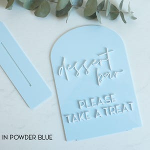 May include: A powder blue arched acrylic sign with white 3D lettering that reads dessert bar and PLEASE TAKE A TREAT. The set includes a matching rectangular base with a slot for the sign to stand upright, featuring a smooth finish.