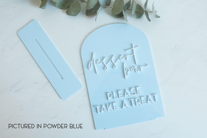 May include: A powder blue arched acrylic sign with white 3D lettering that reads dessert bar and PLEASE TAKE A TREAT. The set includes a matching rectangular base with a slot for the sign to stand upright, featuring a smooth finish.