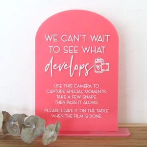 May include: A pink acrylic sign with an arched top. White text reads "WE CAN'T WAIT TO SEE WHAT develops" with a film roll graphic. Additional text instructs users to capture moments and pass the camera along. A small base supports the sign.