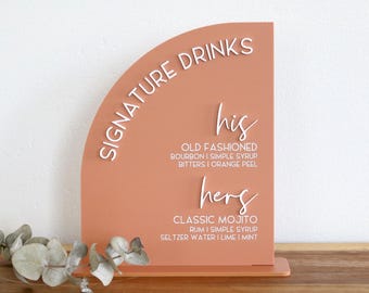 May include: A coral-colored acrylic sign with the words "SIGNATURE DRINKS" in white script. The sign lists cocktail ingredients for "his" and "hers" drinks, including an Old Fashioned and a Classic Mojito. The sign is on a wooden surface.