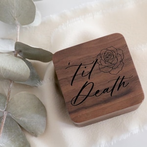 May include: A square, wooden box with a rose illustration and the words "Til Death" in elegant script. The box is a rich brown color and sits on a white, textured surface. Eucalyptus leaves are in the background.