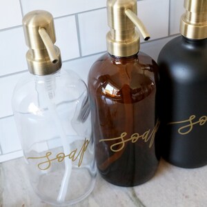 Pu&ograve; includere: Tre dispenser di sapone in vetro con pompe dorate ed etichette che recitano "sapone" in scrittura dorata. I dispenser sono trasparenti, ambrati e neri.
