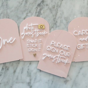 May include: Four blush pink acrylic signs with arched tops. The signs have white text that reads "One", "photo guestbook, snap it, stick it, sign it", "Please sign our guestbook", and "cards and gifts". A gold camera and heart accent the signs.