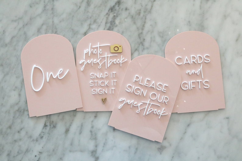 May include: Four blush pink acrylic signs with arched tops. The signs have white text that reads "One", "photo guestbook, snap it, stick it, sign it", "Please sign our guestbook", and "cards and gifts". A gold camera and heart accent the signs.
