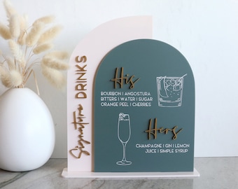 May include: A dual-layered arched sign featuring a pale pink back panel and a dark teal front panel. The sign displays the text "Signature Drinks" in gold, along with "His" and "Hers" headings, cocktail illustrations, and ingredient lists.