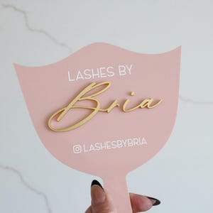 May include: A pale pink handheld sign with a wavy top edge featuring the white text LASHES BY and the Instagram handle LASHESBYBRIA. The name Bria is displayed in a raised gold script font centered on the surface of the pink sign.