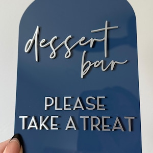 May include: A navy blue acrylic sign with a metallic silver finish. The sign reads "dessert bar" in cursive and "PLEASE TAKE A TREAT" in block letters. The top of the sign is arched.