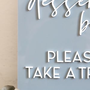 May include: A light blue rectangular sign featuring white 3D lettering. The top text reads dessert bar in a cursive script, while the bottom text reads PLEASE TAKE A TREAT in a clean, sans-serif font. The sign has a smooth, matte finish.