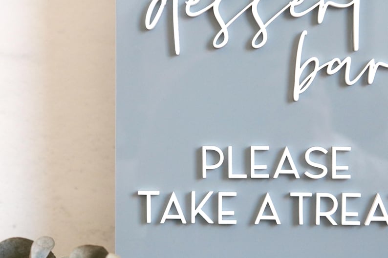 May include: A light blue rectangular sign featuring white 3D lettering. The top text reads dessert bar in a cursive script, while the bottom text reads PLEASE TAKE A TREAT in a clean, sans-serif font. The sign has a smooth, matte finish.