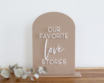 May include: An arched, light brown sign featuring the white, raised text "OUR FAVORITE love STORIES" in a combination of sans-serif and script fonts. The sign is mounted on a rectangular base and is positioned next to a sprig of green eucalyptus.
