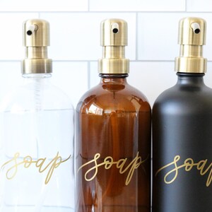 Pu&ograve; includere: Tre dispenser di sapone in vetro con pompe dorate e scritta dorata che dice "sapone" su ogni bottiglia. Le bottiglie sono trasparenti, ambrate e nere.