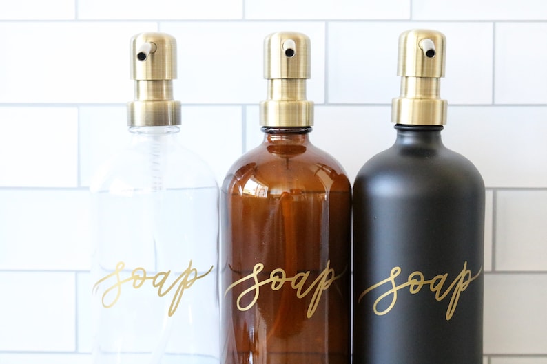 Pu&ograve; includere: Tre dispenser di sapone in vetro con pompe dorate e scritta dorata che dice "sapone" su ogni bottiglia. Le bottiglie sono trasparenti, ambrate e nere.