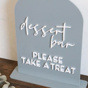 May include: An arched, light gray sign with white 3D lettering. The top text reads "dessert bar" in a cursive script, and the bottom text reads "PLEASE TAKE A TREAT" in a bold, sans-serif font. The sign is mounted on a rectangular base.