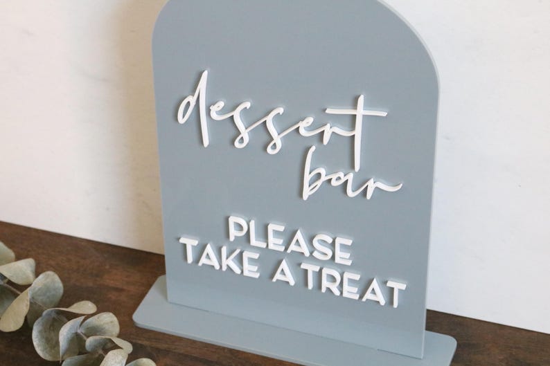 May include: An arched, light gray sign with white 3D lettering. The top text reads "dessert bar" in a cursive script, and the bottom text reads "PLEASE TAKE A TREAT" in a bold, sans-serif font. The sign is mounted on a rectangular base.