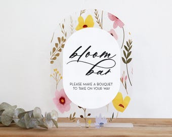 May include: An oval-shaped sign with a floral design and the words "bloom bar" in black script. The sign has a white oval in the center with the text "PLEASE MAKE A BOUQUET TO TAKE ON YOUR WAY". The background features pink and yellow flowers.