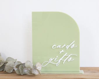 May include: A sage green acrylic arch sign featuring the white text cards + gifts in a cursive script font. The sign has a flat base and a smooth, matte finish, standing upright as a display piece for events or special occasions.