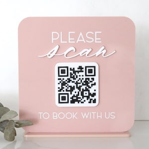 May include: A pink sign with rounded corners features the words "PLEASE scan TO BOOK WITH US" in white lettering. A QR code is in the center. The sign is on a clear stand.