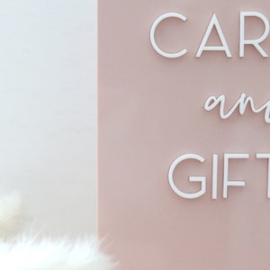 May include: A sign with the words "CARDS and GIFTS" in white, against a pale pink background. The sign is likely for a wedding or event, with a soft, elegant aesthetic.