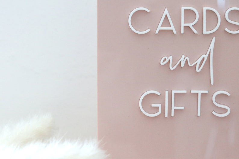 May include: A sign with the words "CARDS and GIFTS" in white, against a pale pink background. The sign is likely for a wedding or event, with a soft, elegant aesthetic.