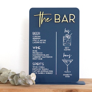 May include: A rectangular navy blue bar menu sign with rounded corners featuring gold 3D lettering that reads The BAR. The sign lists beer, wine, and spirits options in white text, alongside white line drawings of a dirty martini and amaretto sour.